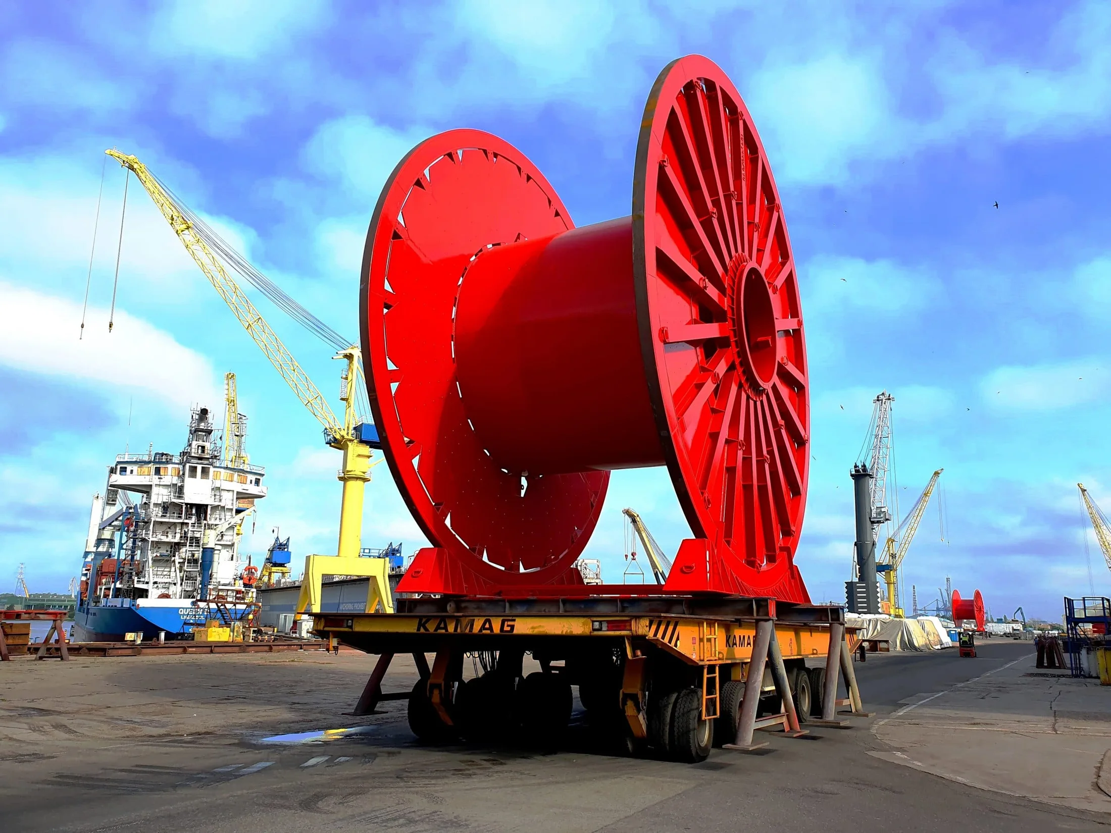 Carretel vermelho sobre transportador no cais, com navio ao fundo